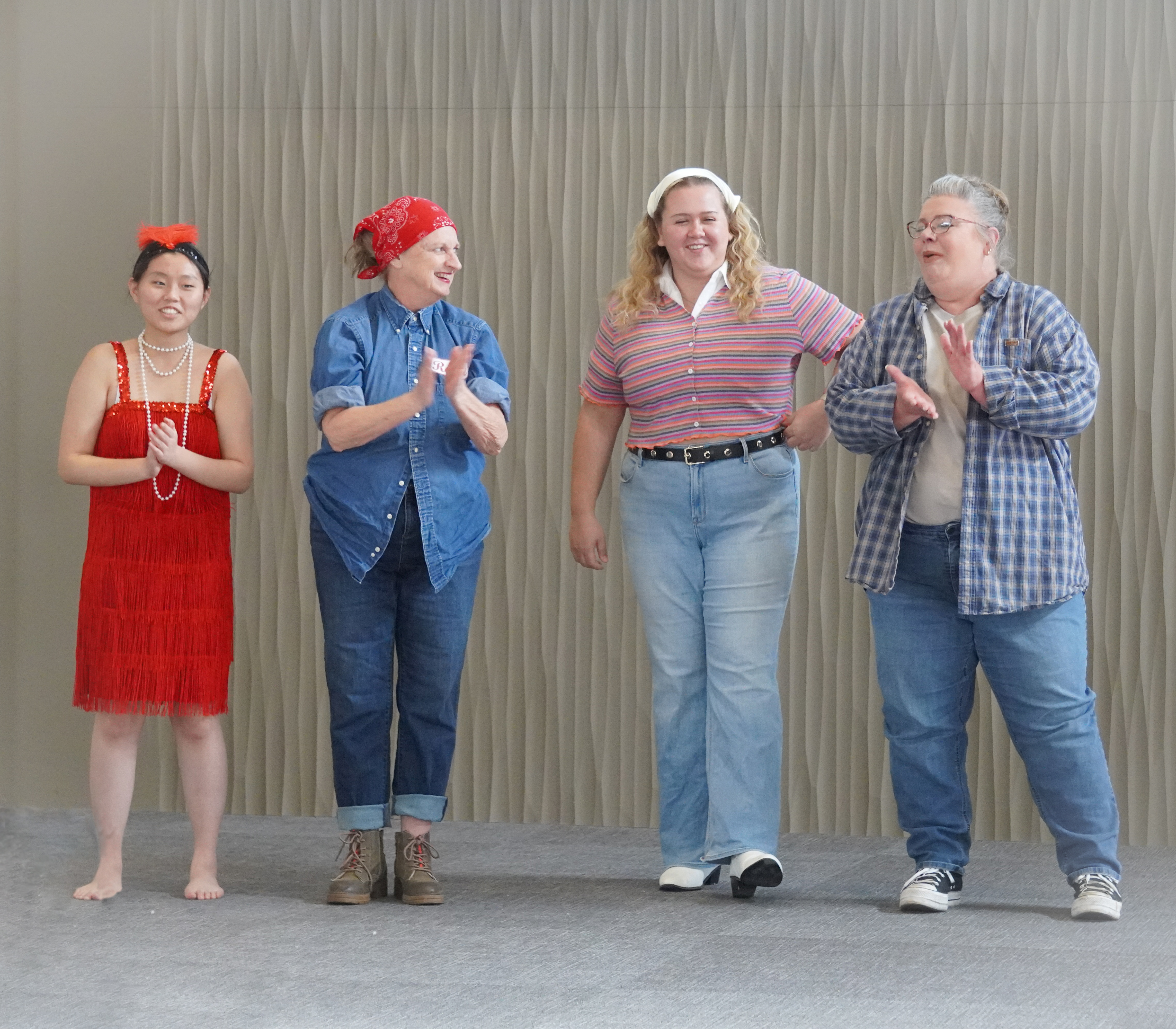 Four female models standing side by side with each other. The first female wears a flapper dress and accessories. The next female is dressed as Rosie the Riveter. The next female wears a hippie-inspired look. The fourth female wears a grunge era inspired outfit. 